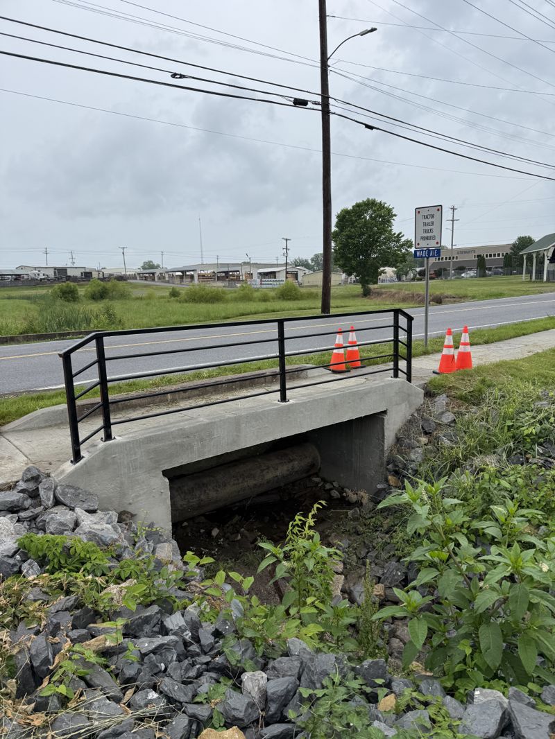 Black steel pipe handrail fabricated and installed along a concrete culvert headwall beside a commercial roadway, with traffic cones set out on the shoulder