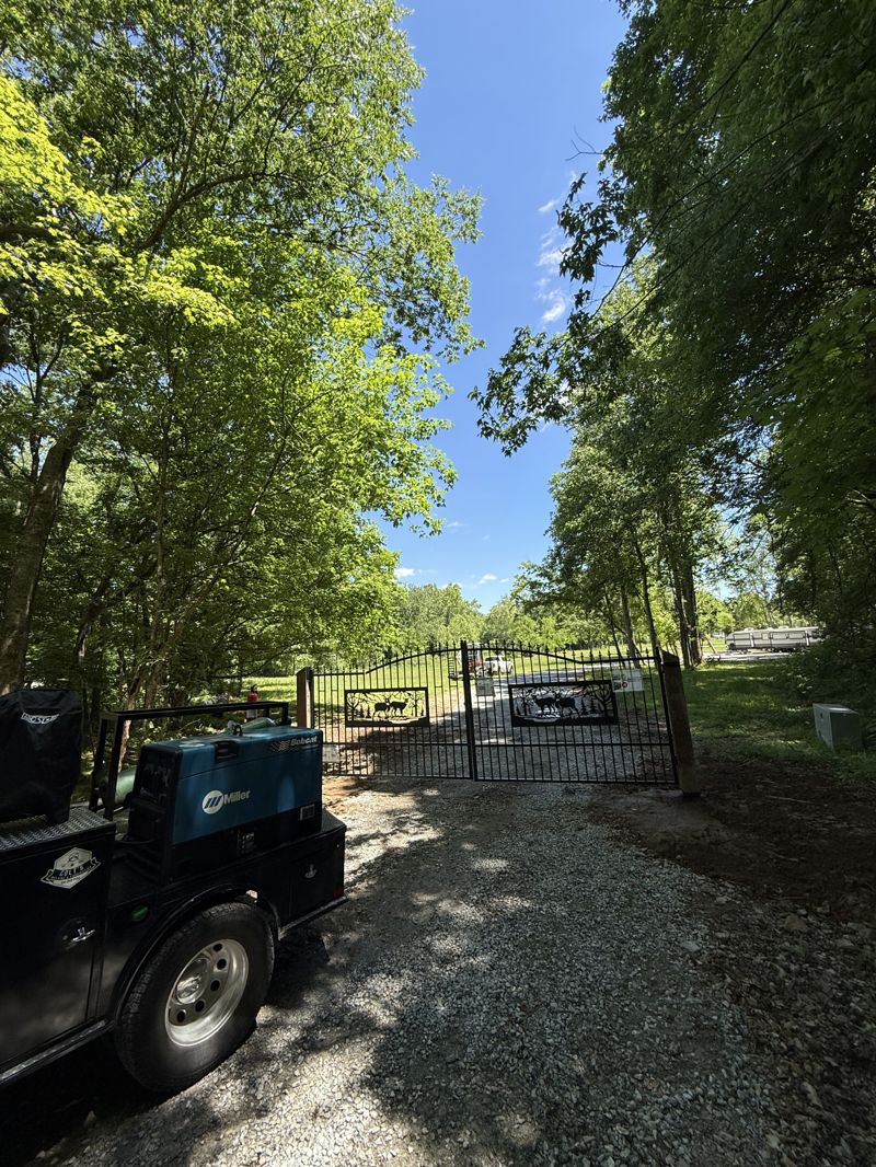 Custom double-swing steel driveway gate with an arched top, vertical pickets, and decorative cut-metal wildlife panels, installed on timber posts across a gravel drive with a mobile welding truck parked alongside
