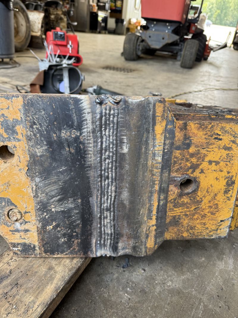 Heavy steel reinforcement plate welded onto the yellow-painted frame of a piece of construction equipment, showing rows of multi-pass stringer weld beads across the repaired joint