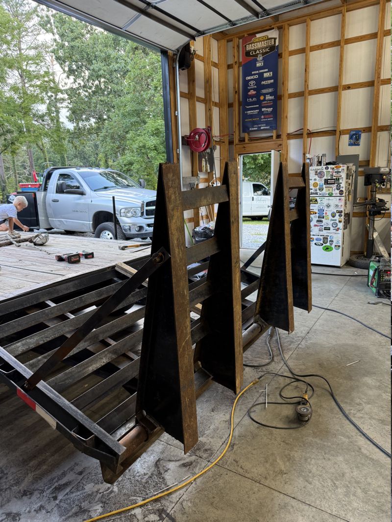 Equipment trailer in the shop with its deck boards removed to expose the steel frame and crossmembers, and newly fabricated stand-up steel ramps welded to the rear